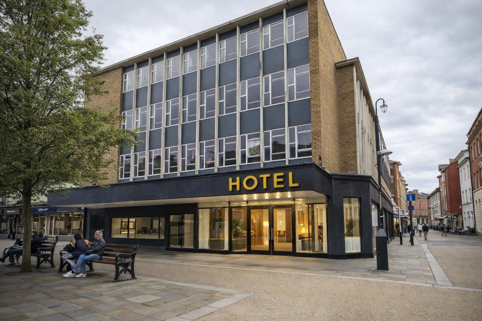 Former Debenhams building on Worcester High Street reimagined as a modern hotel, with a lit entrance, large windows, and people sitting on benches nearby.