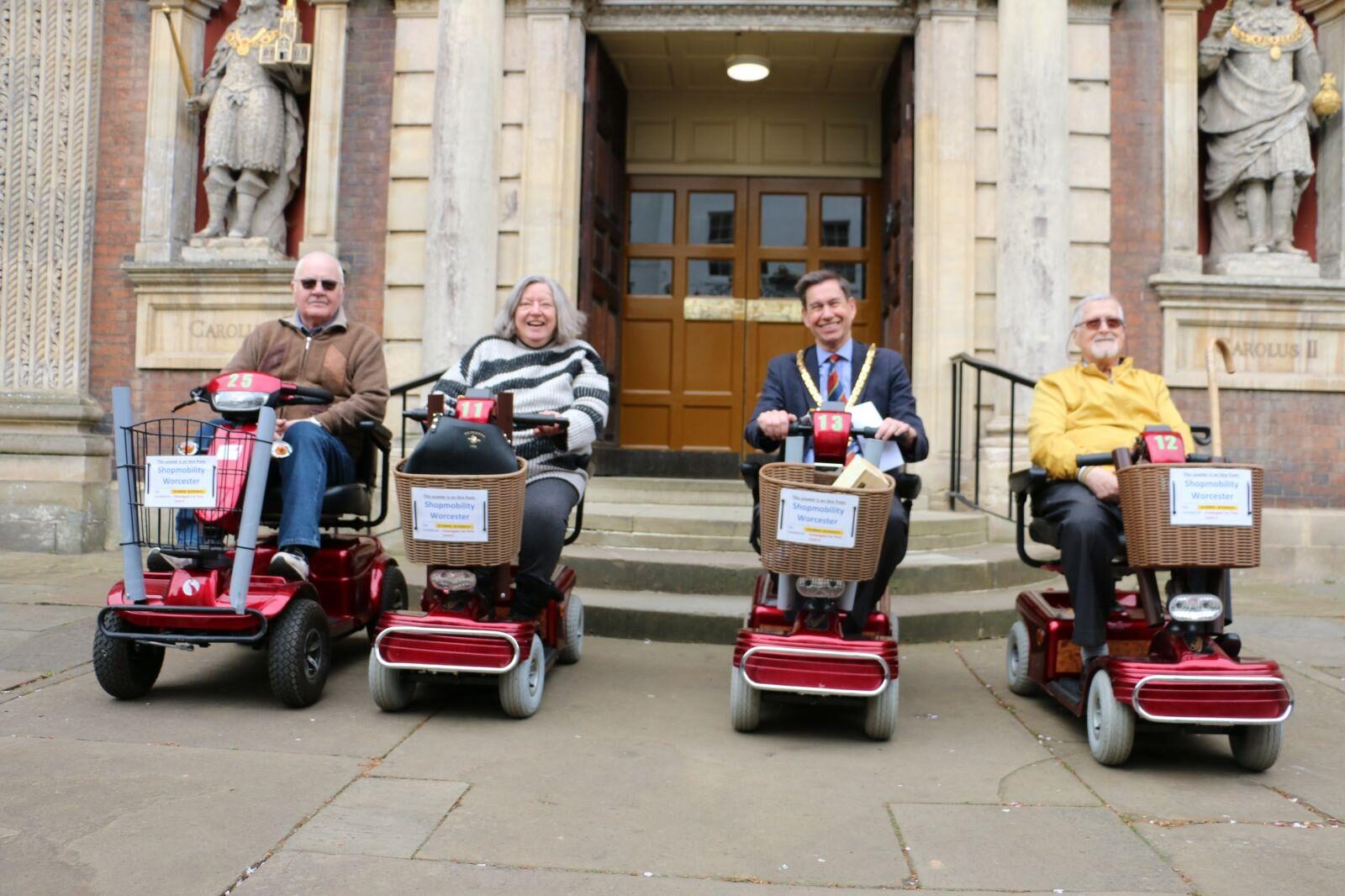 The Mayor of Worcester and three others sit on mobility scooters in front of a historic civic building, demonstrating accessible transport as part of a ShopMobility Worcester visit.