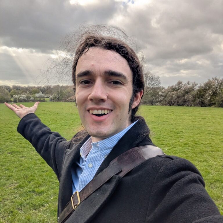 Ed Kimberley smiling and gesturing towards an open green field under a cloudy sky, with sunlight breaking through in the distance.